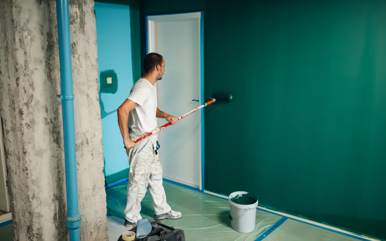 A painter rolling one wall of a room with a masked door, trim and floor around them — the separate surfaces a quote should price individually.